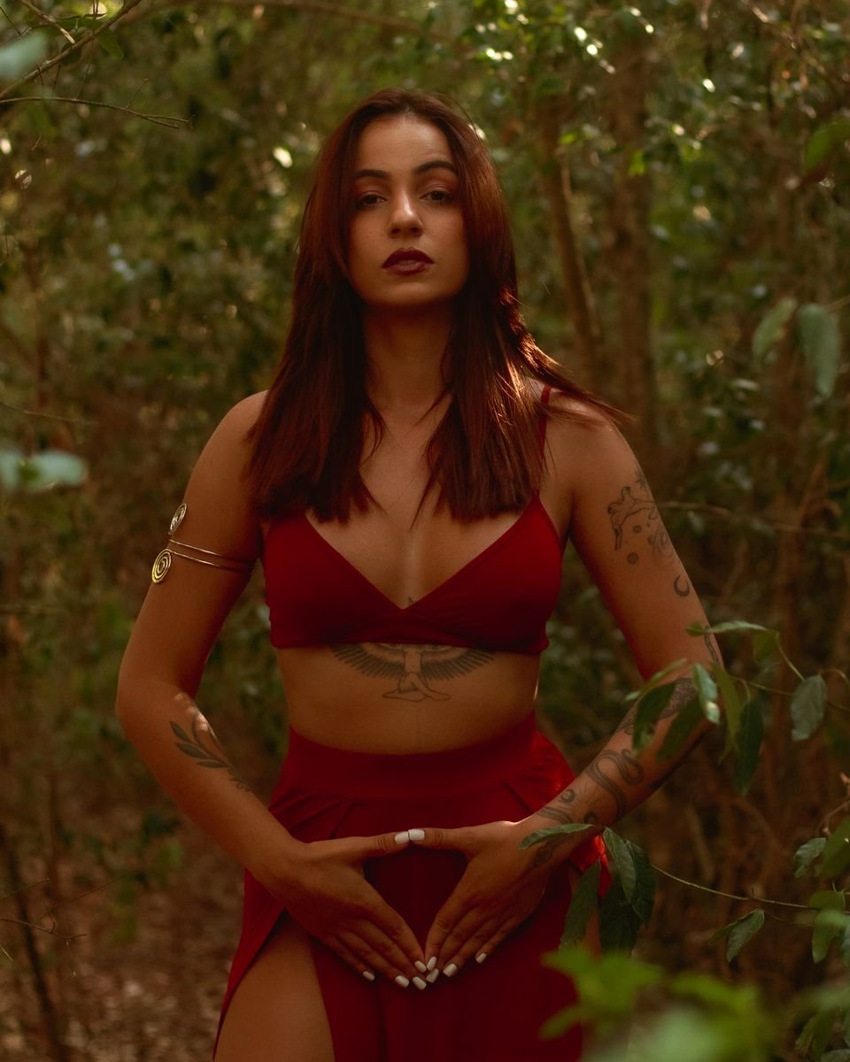 Mulher de vermelho na floresta, com as mãos em forma de triângulo sobre o ventre